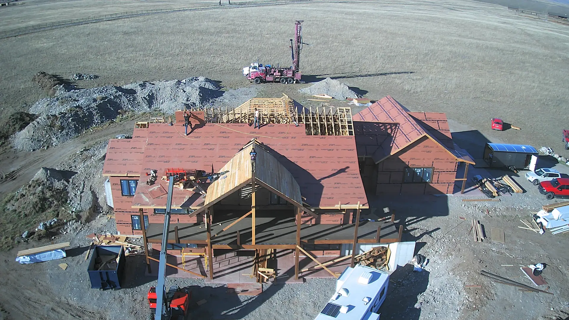 A drone view of a large home mid-build, roof underlayment going on while a well rig works beside it