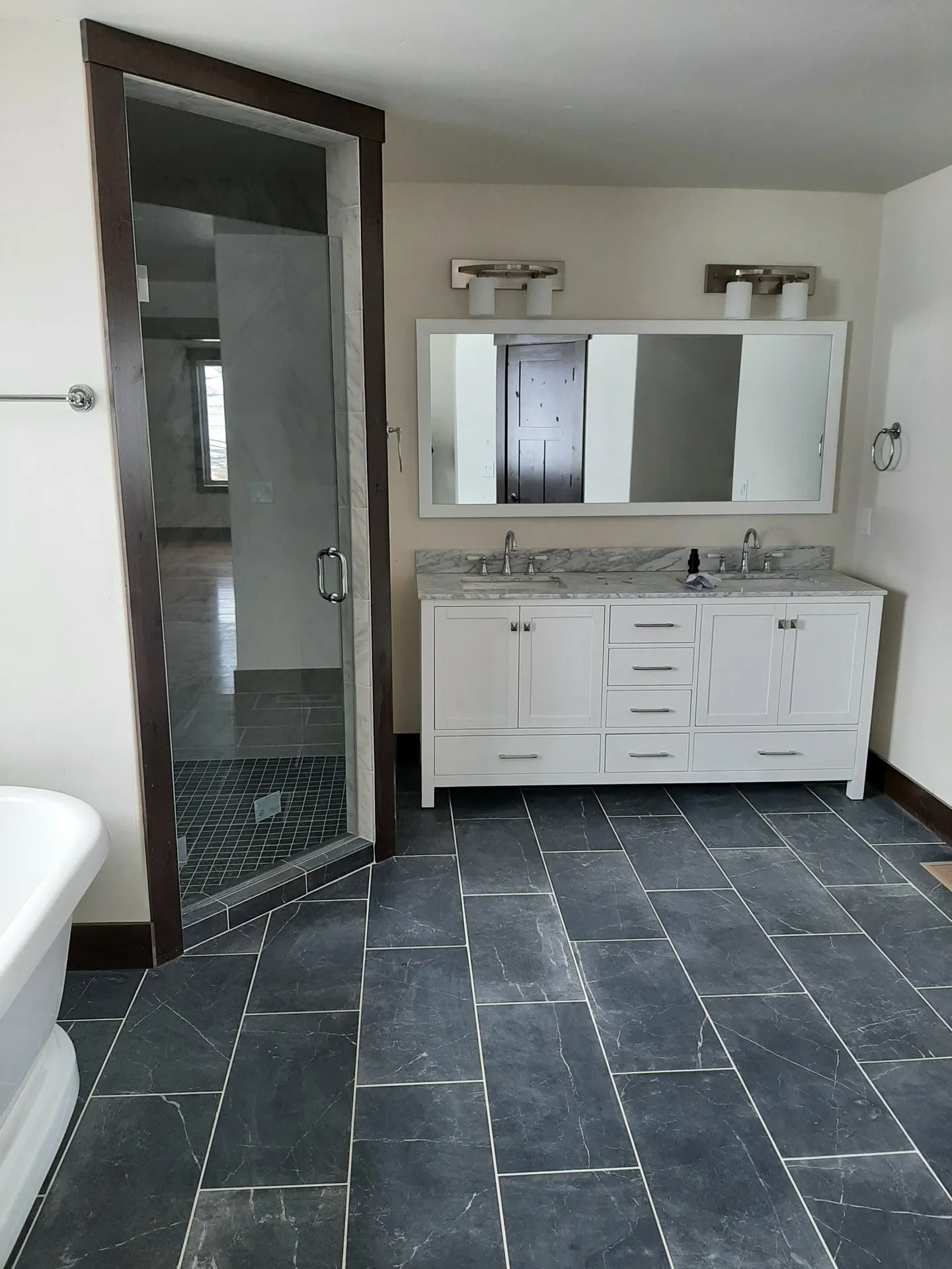 A bathroom with a white double vanity, a tiled glass shower and a dark slate-tile floor