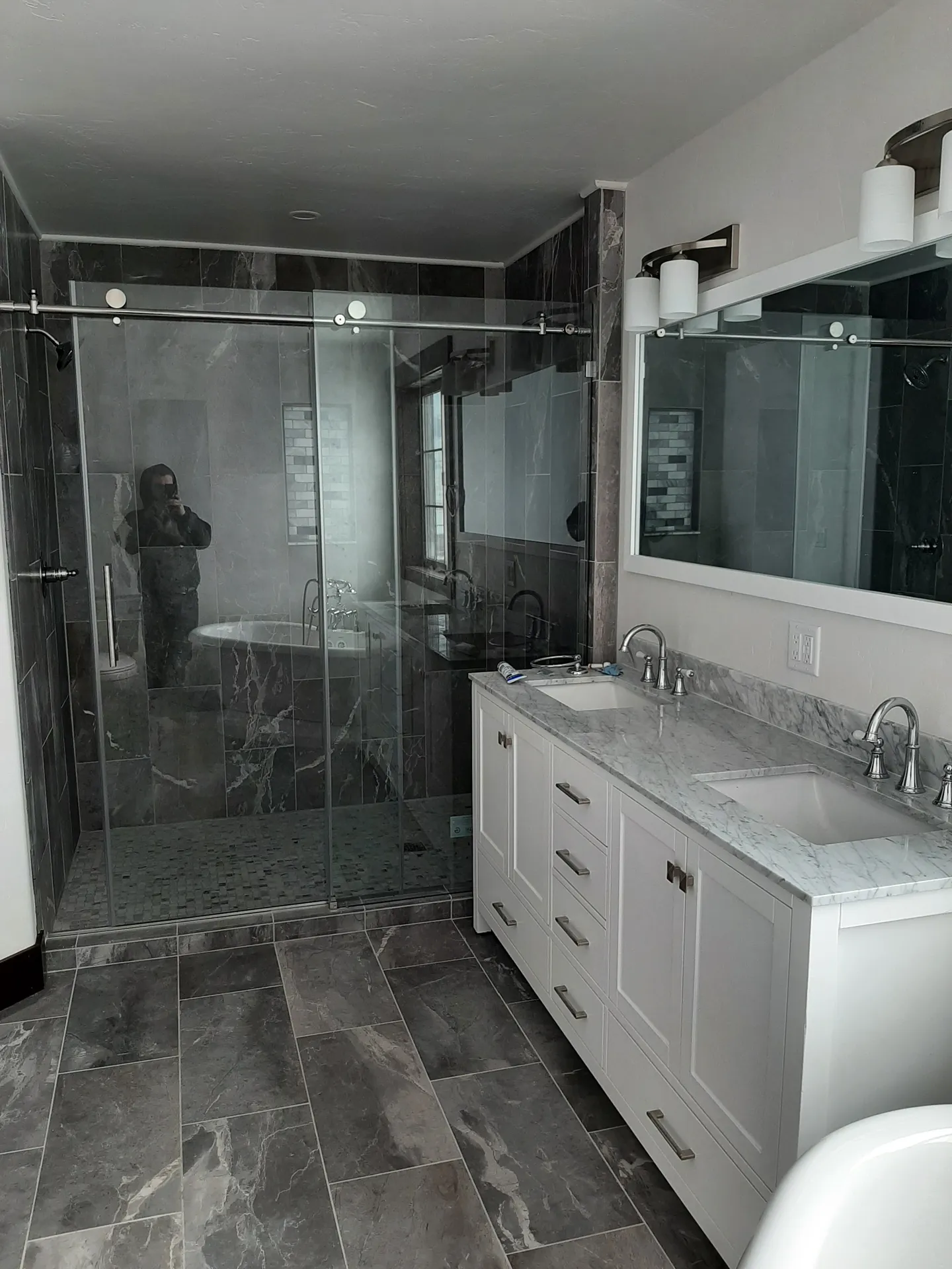 A frameless glass shower in marbled grey tile beside a white vanity with a stone top