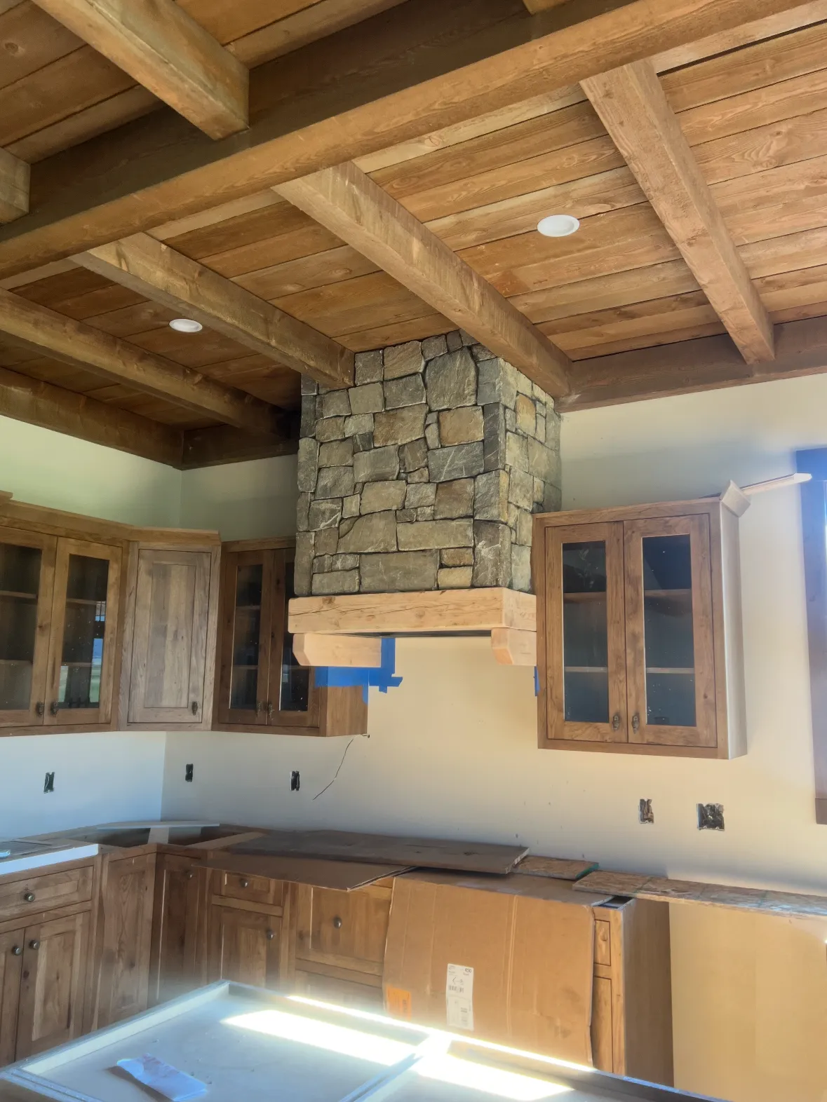 Wood kitchen cabinets going in under a timber ceiling, a stone-clad range hood above